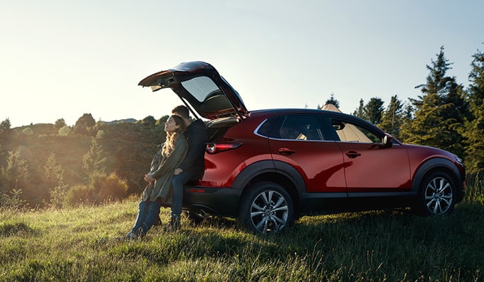 Man and woman sitting against open Mazda CX-30 boot in countryside. 